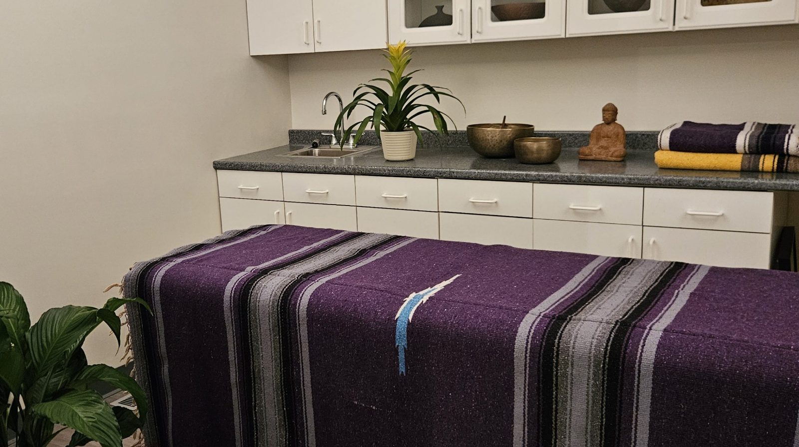 The Studio: a massage table covered with a purple striped blanket in front of white cabinets, a sink and a small Buddha statue